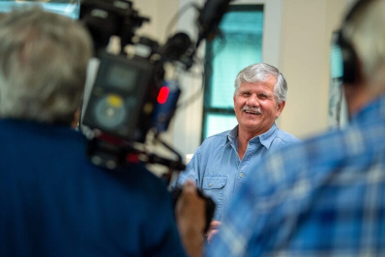 In a cozy room, an older man with gray hair and a mustache smiles while speaking. Dressed in a blue shirt, he shares his insights as the cameraman in blue flannel captures every moment.