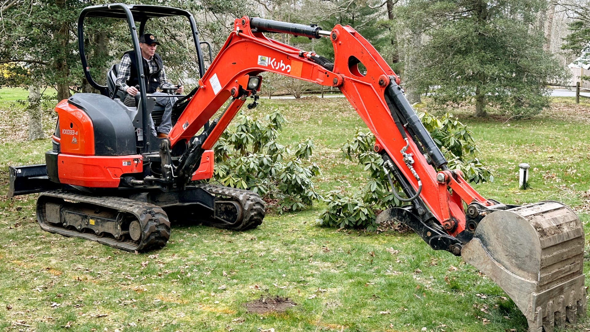 A person operates a small orange excavator with a digging bucket in a grassy area. Nearby, there are some bushes and trees. The ground appears disturbed, as if prepared for construction or landscaping.