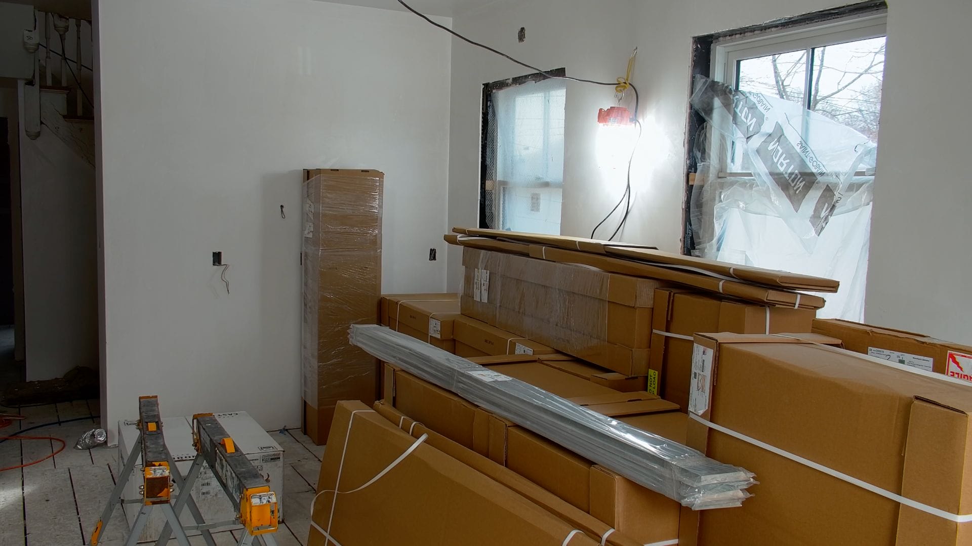 A room under renovation with stacked cardboard boxes, ladders, and plastic-wrapped materials. Walls and windows are partially covered, indicating ongoing construction work. A light illuminates the space, and the floor is unfinished.