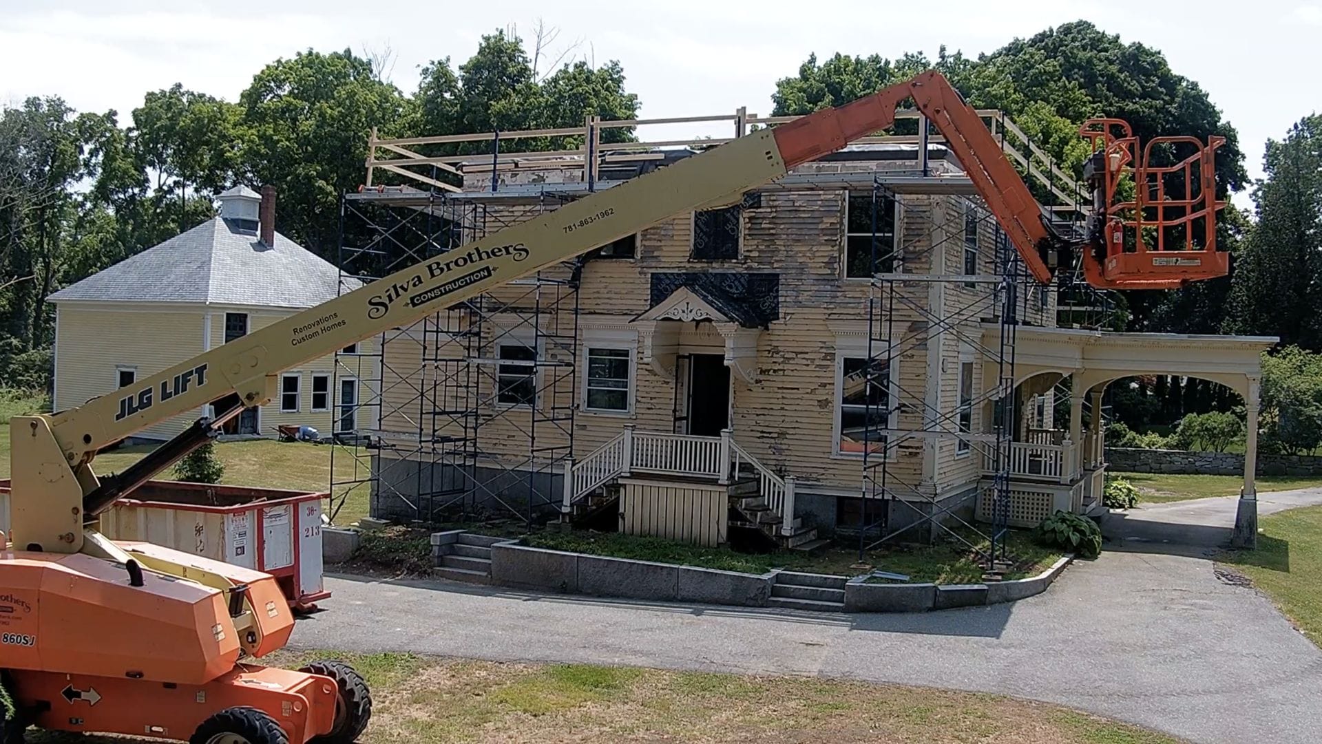 A vintage two-story house under renovation, surrounded by scaffolding. A boom lift with a basket is positioned at the roof level. Trees and another house can be seen in the background.