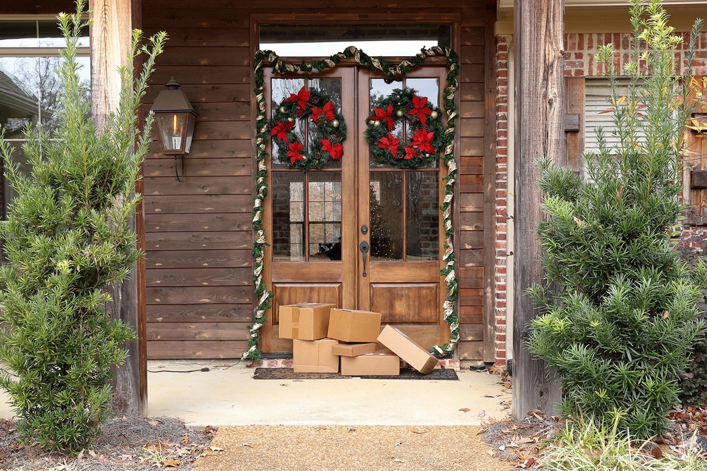Front porch with wooden double doors adorned with red and green holiday wreaths. Several cardboard packages are stacked by the entrance. Two green bushes flank the doorway, and a brick wall is partially visible on the right.